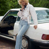 A woman leans against a white vintage Mercedes-Benz car outdoors, wearing a white textured jacket, light blue jeans, and brown loafers. The Mercedes-Benz logo is visible on the car wheel.