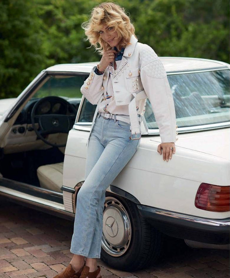 A woman leans against a white vintage Mercedes-Benz car outdoors, wearing a white textured jacket, light blue jeans, and brown loafers. The Mercedes-Benz logo is visible on the car wheel.
