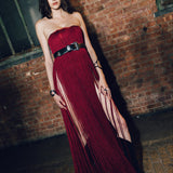 A woman stands in a brick warehouse wearing a strapless deep red fringe gown with a wide black belt and black pointed shoes.