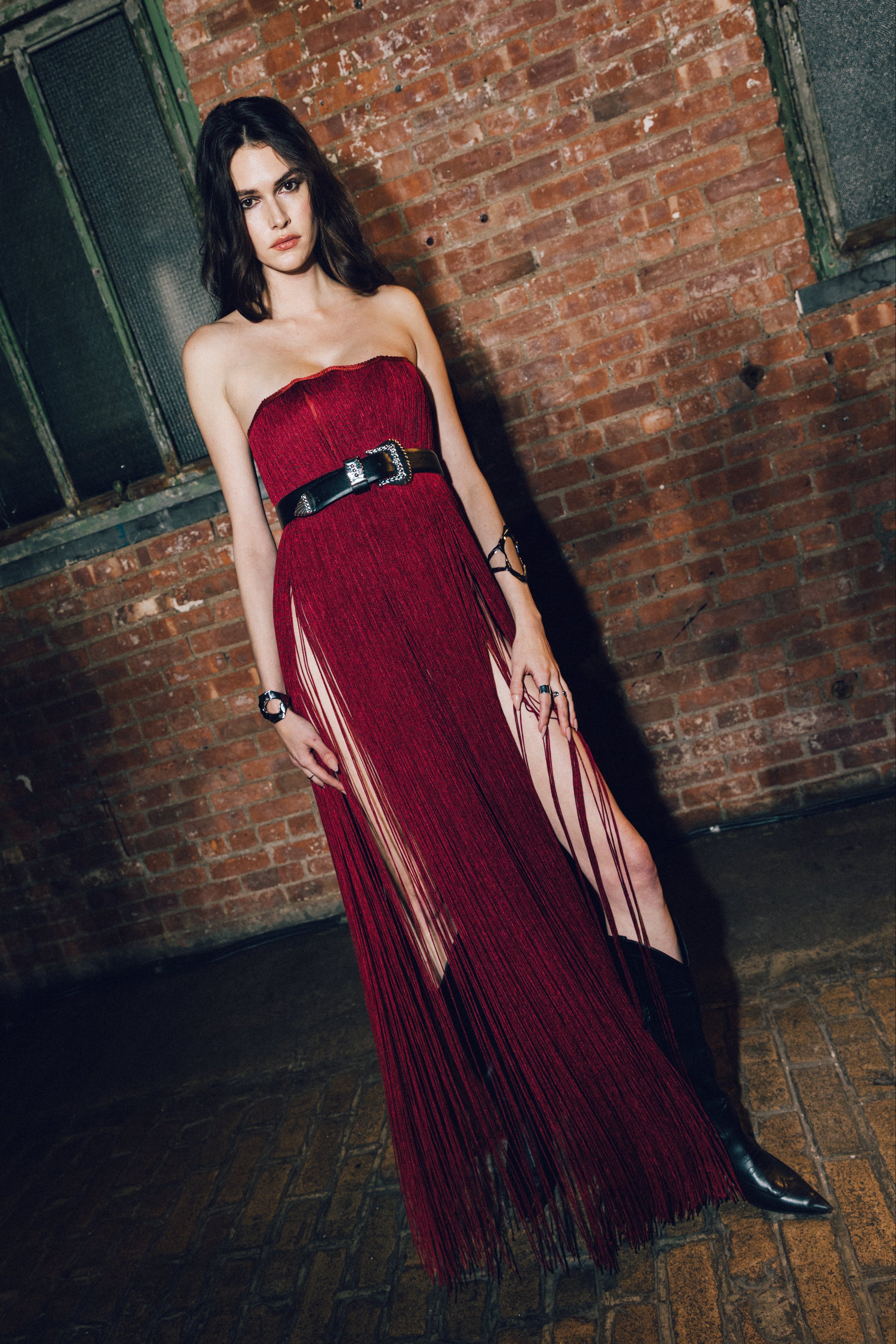 A woman stands in a brick warehouse wearing a strapless deep red fringe gown with a wide black belt and black pointed shoes.