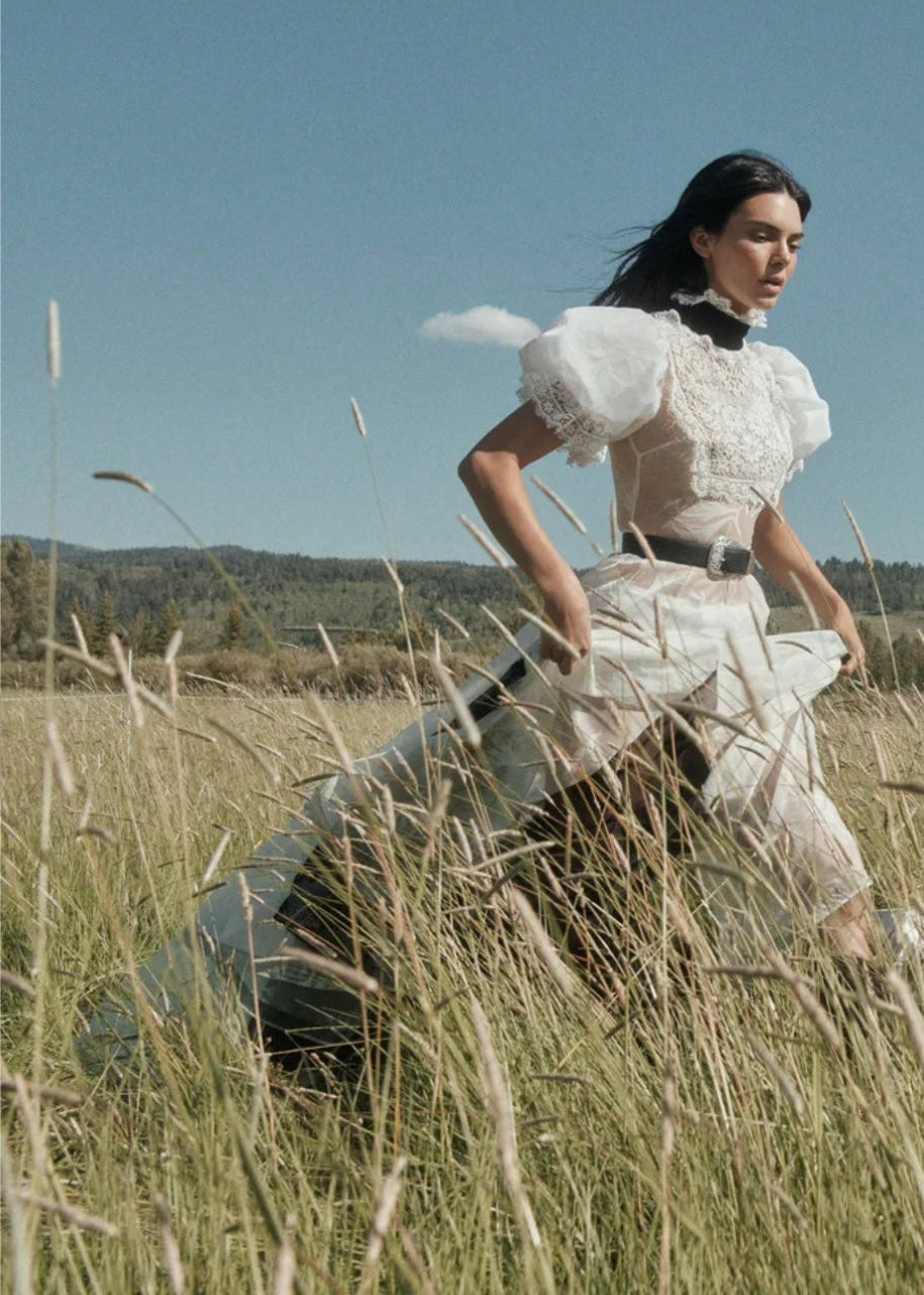 Woman in a white dress walking through tall grass with a suitcase.