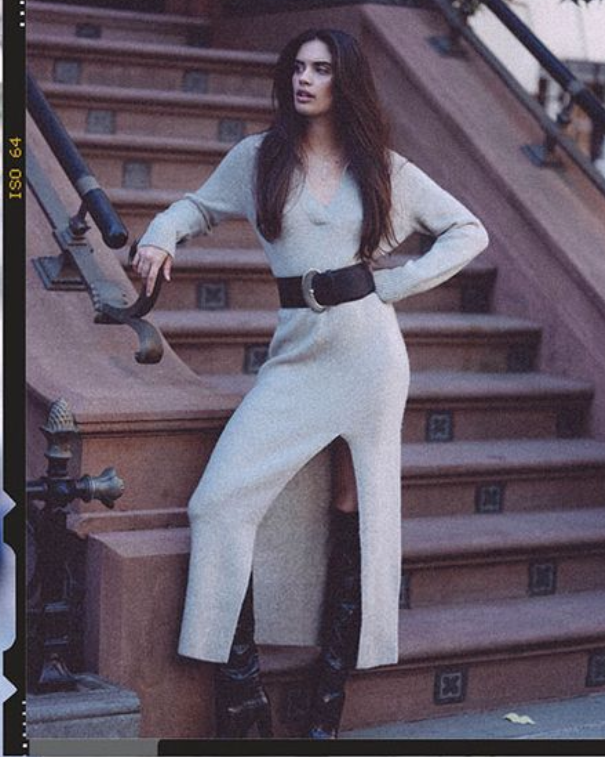 A woman wearing a light beige knit long-sleeve dress with a high slit and a wide black belt, posing on outdoor stone steps.