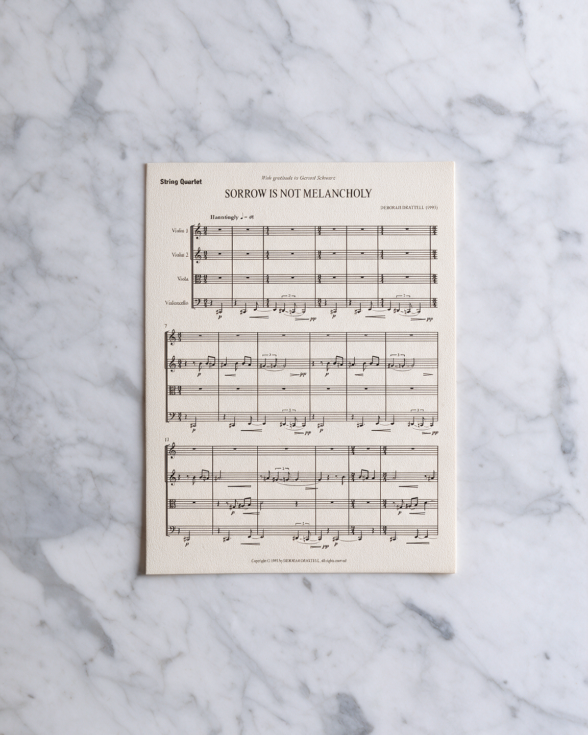 A single sheet of printed sheet music titled 'SORROW IS NOT MELANCHOLY' by Deborah Drattell, placed flat on a white and gray marble surface.