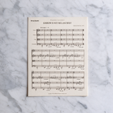 A single sheet of printed sheet music titled 'SORROW IS NOT MELANCHOLY' by Deborah Drattell, placed flat on a white and gray marble surface.